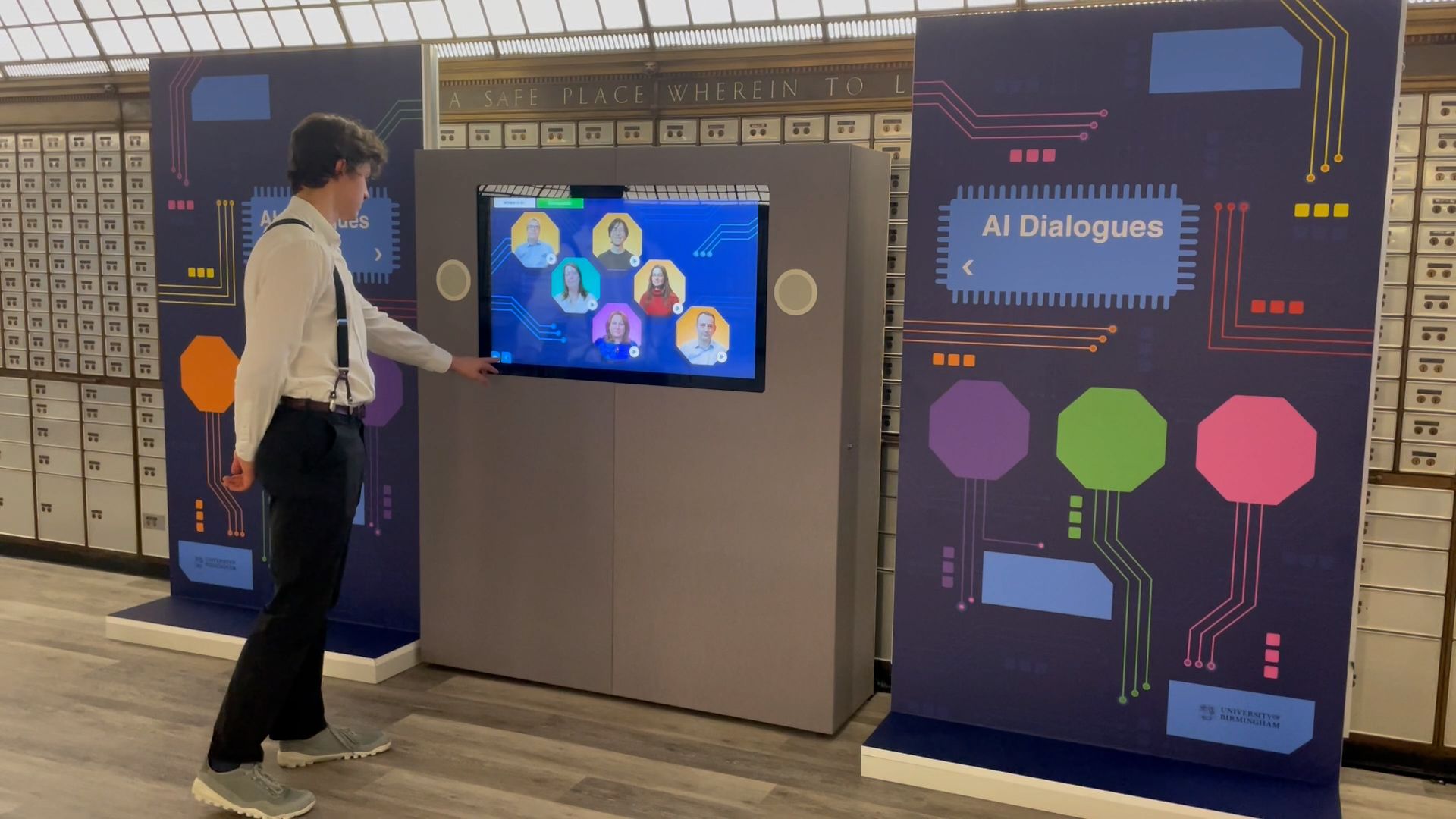 A person in a white shirt and black suspenders interacts with a touchscreen displaying various avatars labeled "AI Dialogues" in a modern setting. The background features a wall of mailboxes and two panels with colorful circuitry designs.