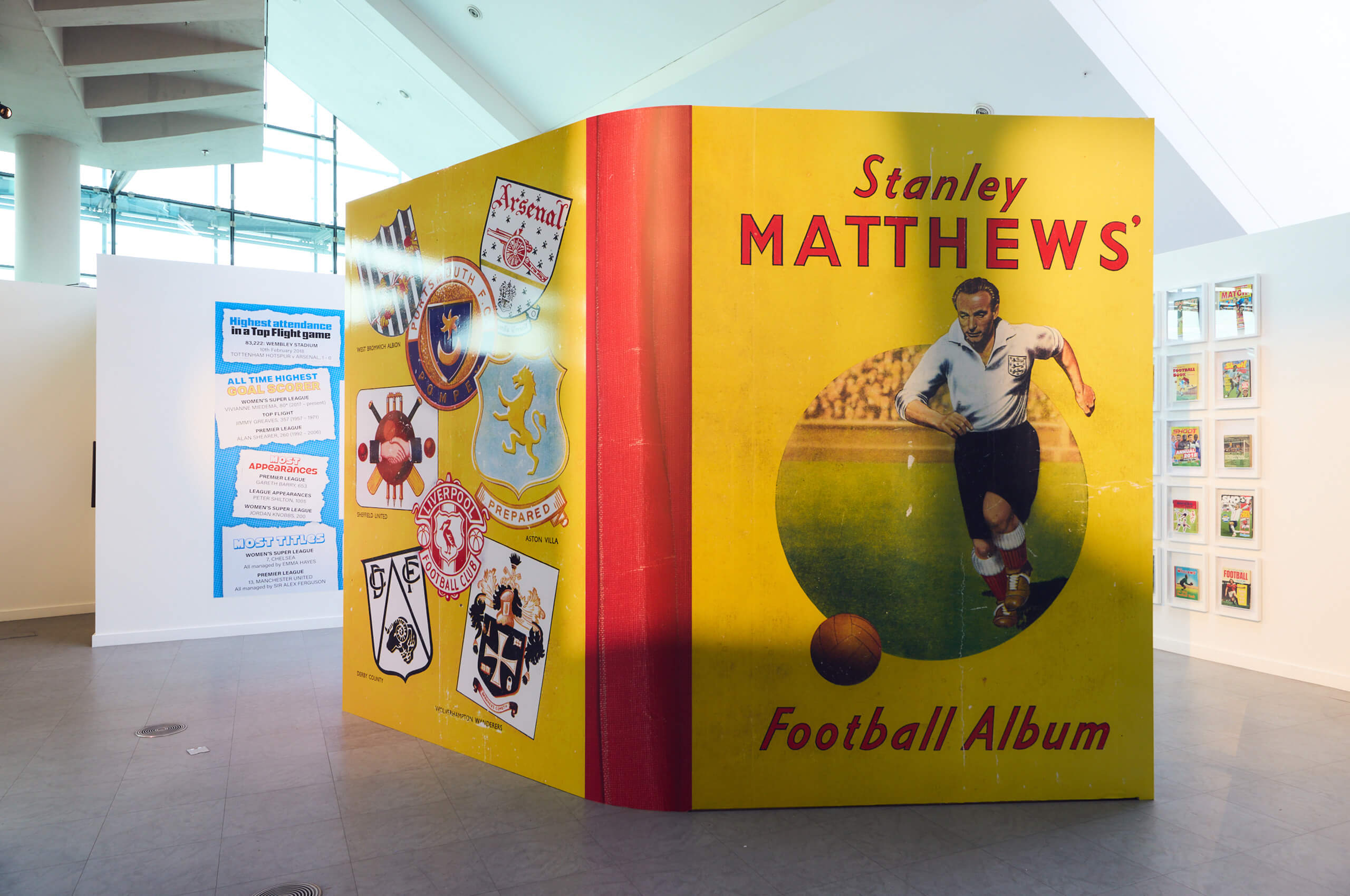 A large display modeled after a vintage football album features an illustration of Stanley Matthews kicking a soccer ball, with team badges and memorabilia on the adjacent panel in a brightly lit exhibition space.