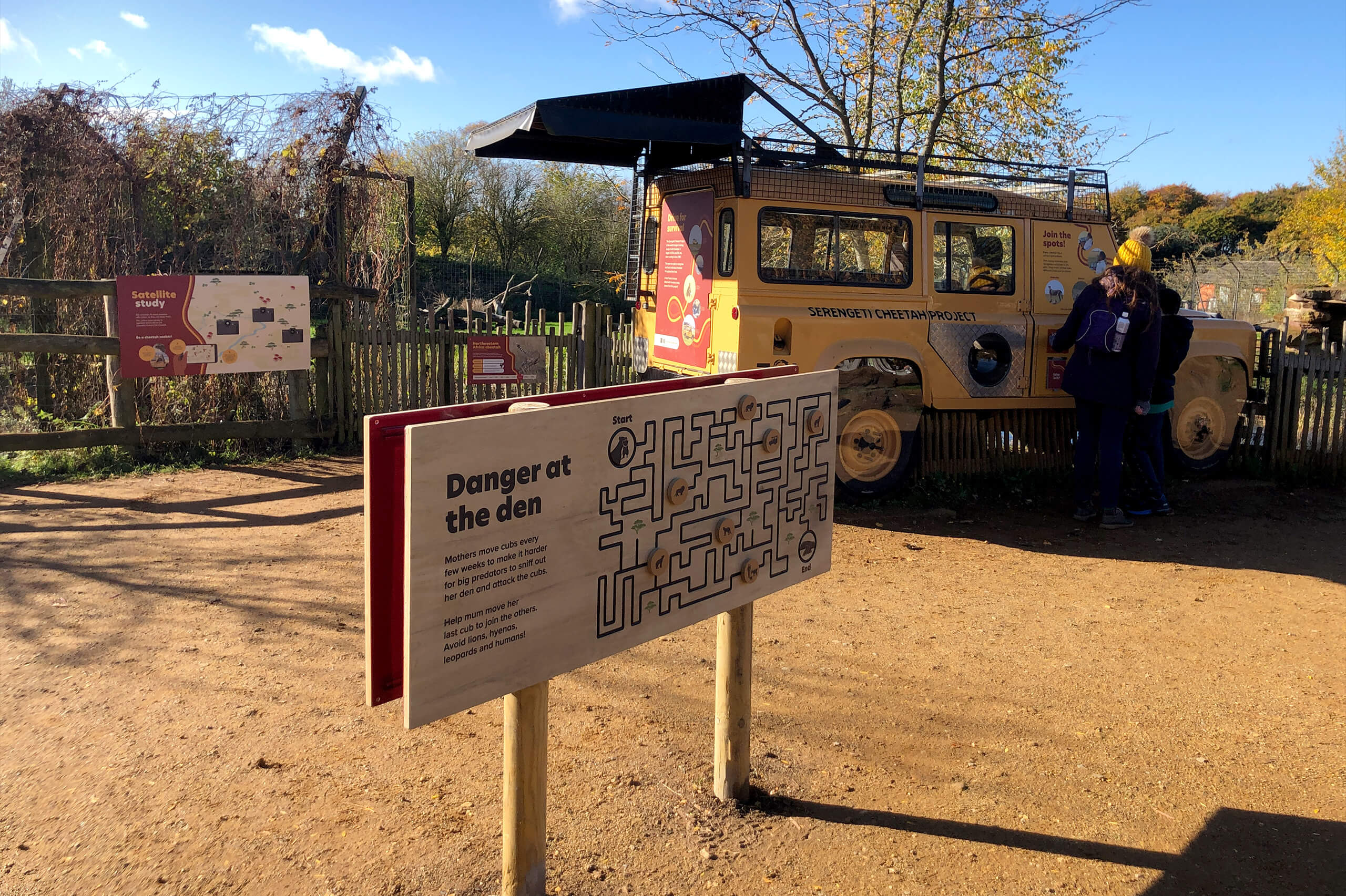 A wooden sign with the text "Danger at the den" and a maze puzzle stands on a dirt path near a yellow safari vehicle, with people and trees in the background.